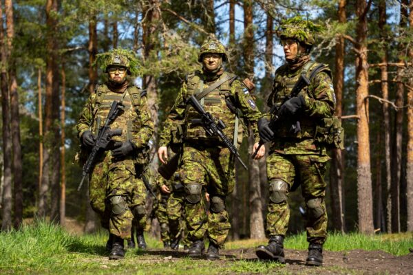 Kolm relvastatud sõdurit digilaigulises vormis ja kiivritega kõnnivad metsarajal; taustal on männimets ning näha on veel üks sõdur nende taga.