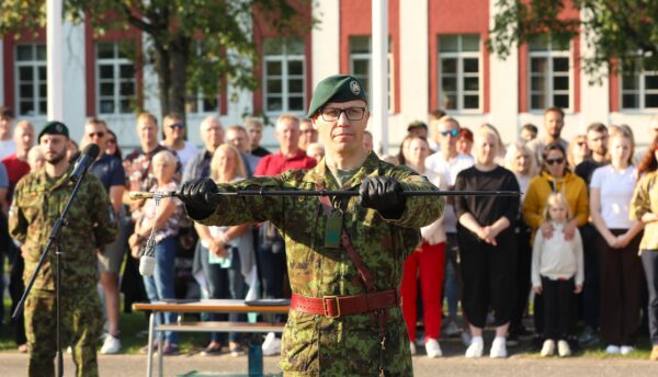 Kaitseväelane digilaigus vormis hoiab pidulikult mõõka vahetustseremoonial, tema taga seisab rivistunud rahvahulk kasarmuhoone ees.