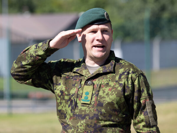 Rohelises digilaigus vormis ja rohelise baretiga kaitseväelane tervitab käega, seistes päikeselisel platsil harjutusväljakul.