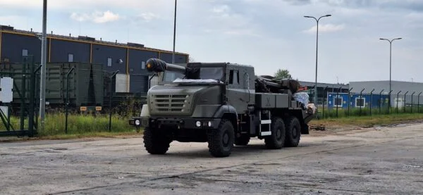 Militaarroheline liikursuurtükk veokipõhisel platvormil seisab tööstusala platsil, toru kaetud kaitsekotiga ja ümberringi laod ning aiad.