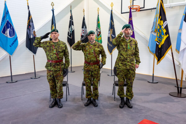 Kolm kaitseväelast digilaigus vormis tervitavad pidulikult siseruumis toimuval tseremoonial, nende taga on üksuste lipud.