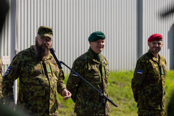 Kolm kaitseväelast digilaigus vormis seisavad õues tseremoonial, üks neist kõneleb mikrofoni ees, teised kuulavad rivis.